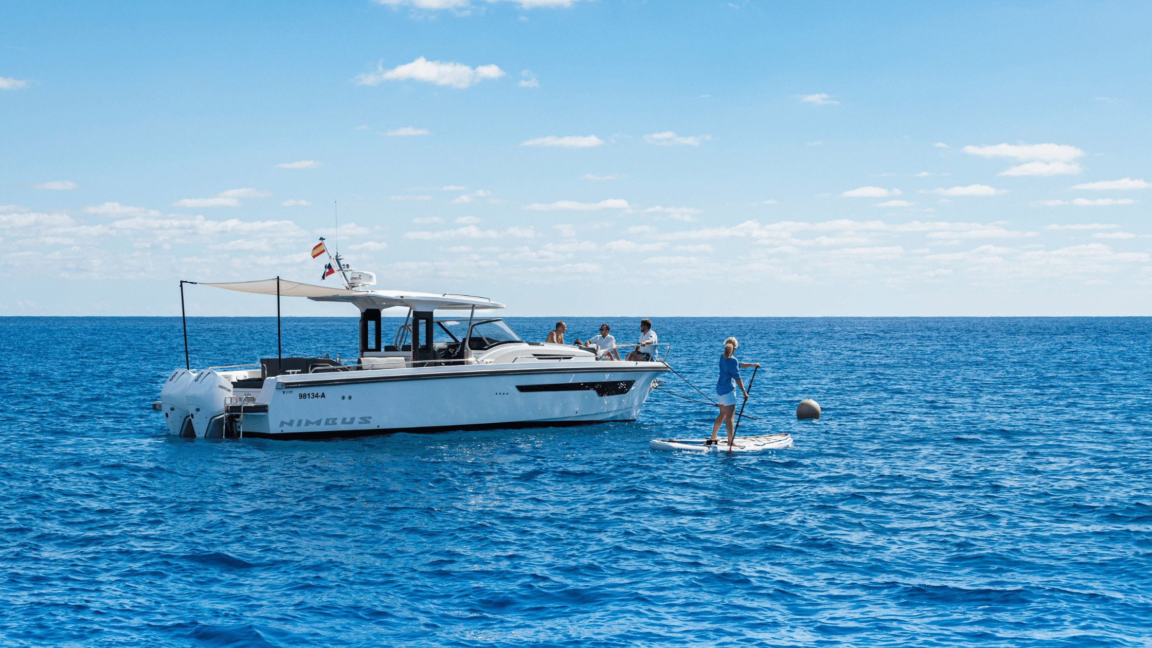 People are lounging on the bow of the Nimbus T11 boat and someone is on a paddle board in the water.